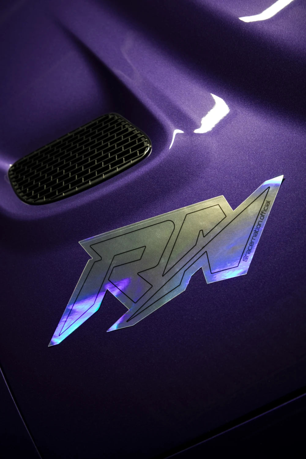 Small holographic Racer Nation RN logo sticker displayed on the bonnet of a purple car, showing its reflective finish.