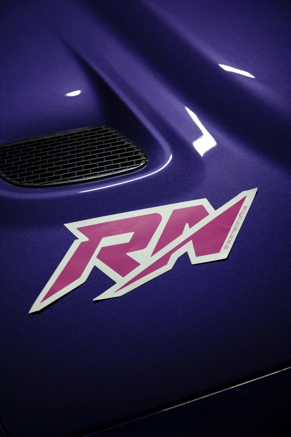 Small pink and white Racer Nation RN logo sticker displayed on the bonnet of a purple car.